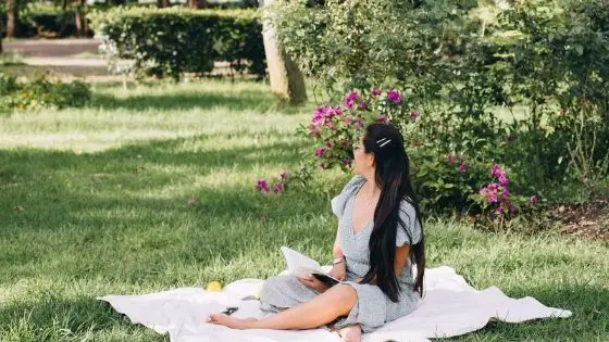 Caucasian girl sitting in a park on a blanket holding a book and looking to the side