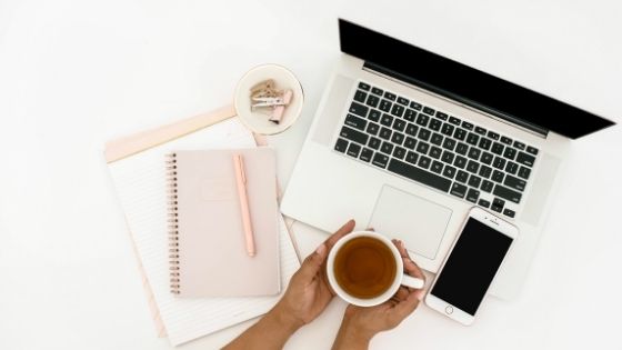 Woman holding cup of tea and desk with laptop, phone and notebooks