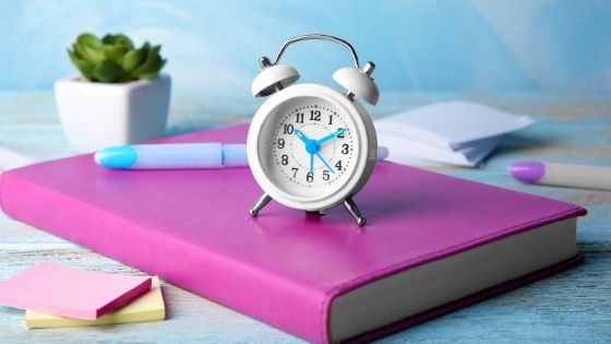 Alarm clock on top of pink notebook on a desk with stationery around it